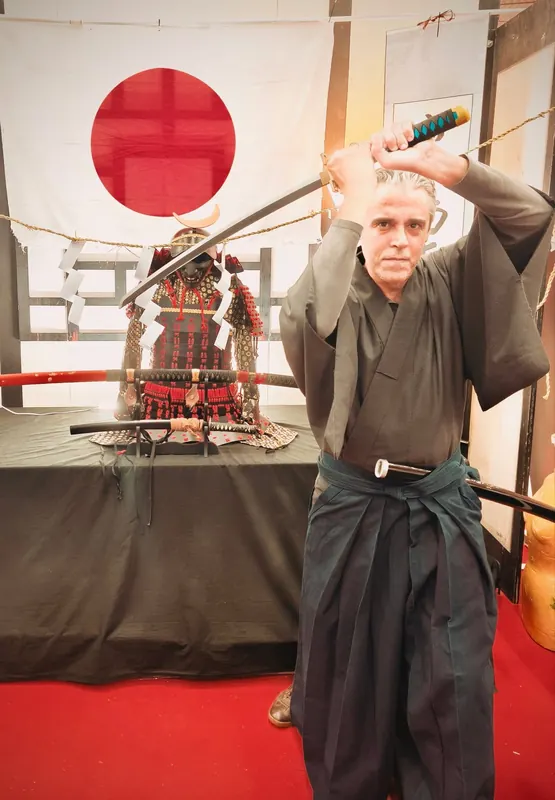 Il maestro in hakama solleva la katana, davanti alla bandiera giapponese e a un'armatura da samurai